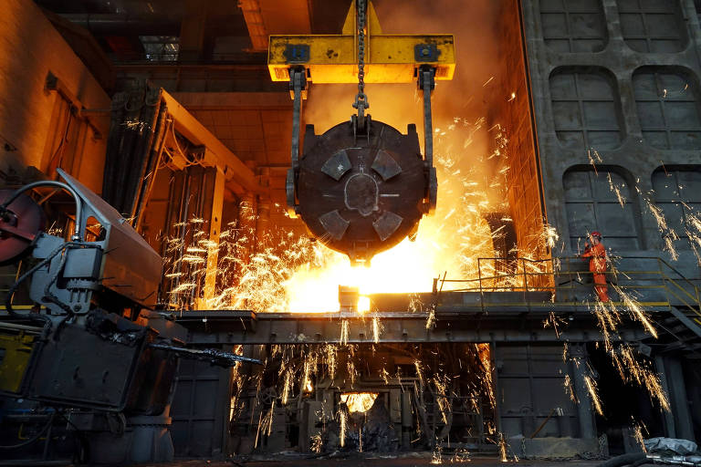 Funcionário (à direita) aparece na entrada de um alto-forno de uma siderúrgica; ao abrir equipamento, saem faíscas do minério de ferro derretido para formar aço