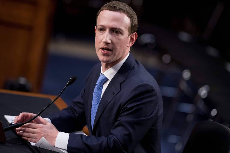 De terno azul marinho e gravata azul, Zuckerberg aparece de perfil esquerdo, olhando para mesa dos senadores (não aparece na imagem), enquanto fala em um microfone de mesa