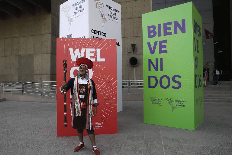 Homem vestido de indígena peruano posa diante de cartaz de boas-vindas da Cúpula das Américas, em Lima