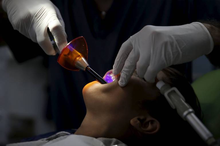 Um dentista usa um instrumento odontológico para tratar uma criança. O aparelho emane uma luz roxa, que pode ser vista dentro da boca do garoto