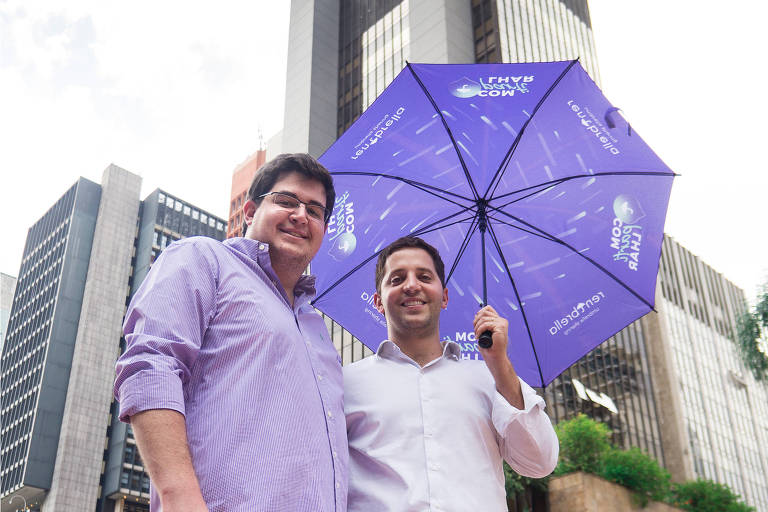 Os empresários Nathan Janovich (esq.) e Freddy Marcos, criadores da Rentbrella, que aluga guarda-chuvas, em avenida em São Paulo, segurando um guarda-chuva roxo da marca