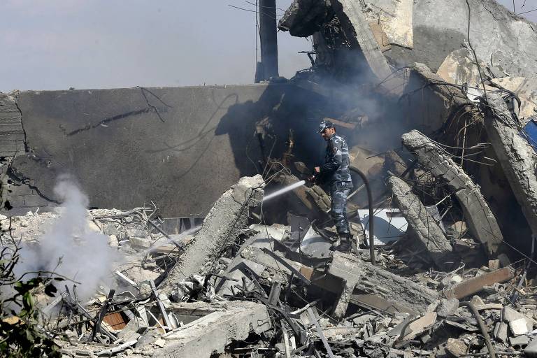 Soldado aparece segurando mangueira de incêndio e jogando água em foco com fumaça entre escombros de prédio; ele aparece ao longe no meio dos destroços