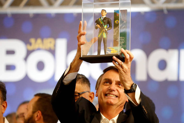Bolsonaro ri, com uma caixa de acrílico com um boneco de si próprio vestido com a faixa presidencial; ao fundo, um banner com o nome do candidato em letras brancas sob fundo azul