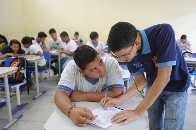 Foto mostra um aluno em pé ensinando colega, sentado, em sala de aula do ensino médio em escola de Jaboatão dos Guararapes, em PE