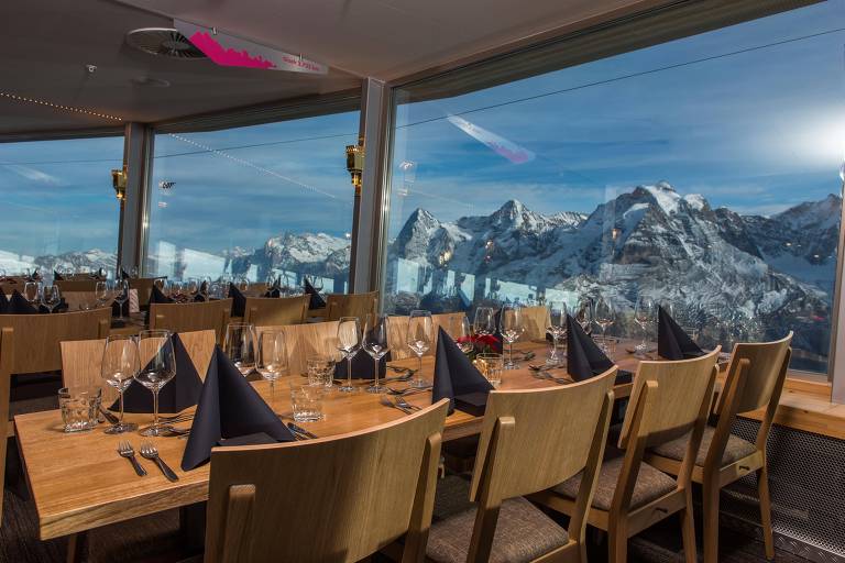 O restaurante 360º tem janelas panorâmicas por todo o seu perímetro. A mesa de oito lugares está com a parte menor encostada na parede da janela. A vista é para as montanhas dos Alpes Suíços