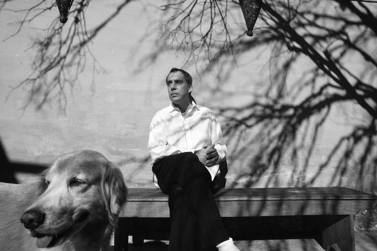 O músico Arnaldo Antunes sentando em um banco; no primeiro plano há um cachorro