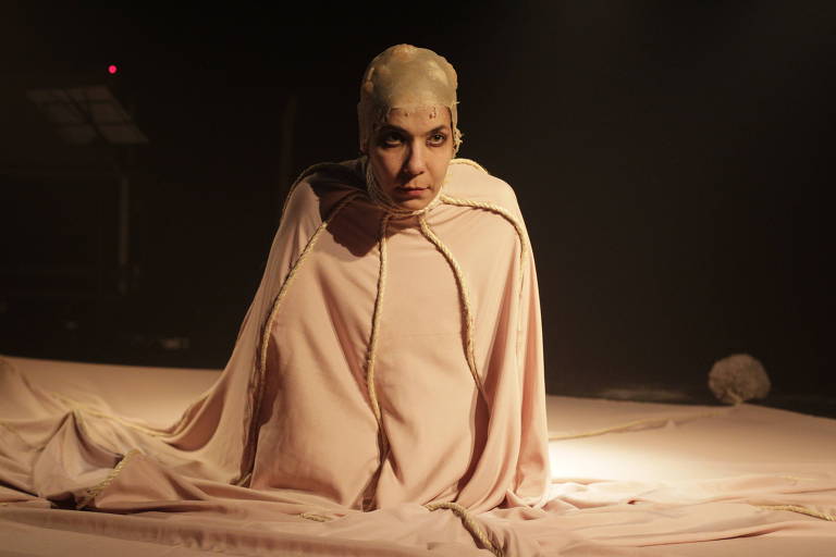 A atriz Fernanda D'Umbra está ajoelhada, coberta por um tecido creme
