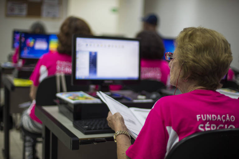Idosa em frente a um computador. Na sala é possível ver outros alunos