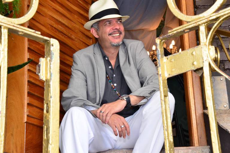 O sambista Marcus Marmello está sentado; ele sorri e usa um blaser, uma calça e um chapéu