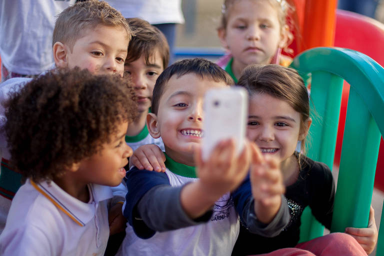 Seis crianças, de 3 a 4 anos, brincam com um celular em meio ao parquinho de uma creche. Algumas delas estão sorrindo, enquanto outras parecem mais desanimadas. Todas vestem uniforme.