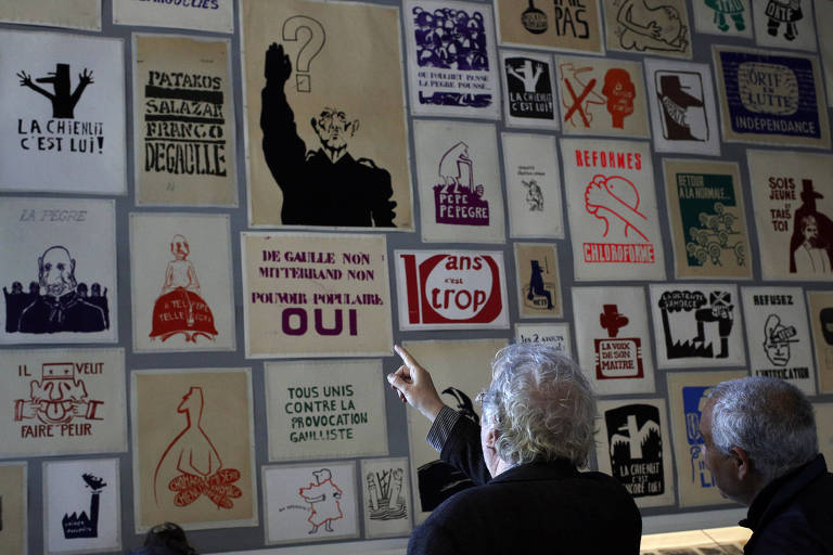 Homem de cabelo branco aponta para série de cartazes com diferentes mensagens em exposição; ao lado dele, outro homem