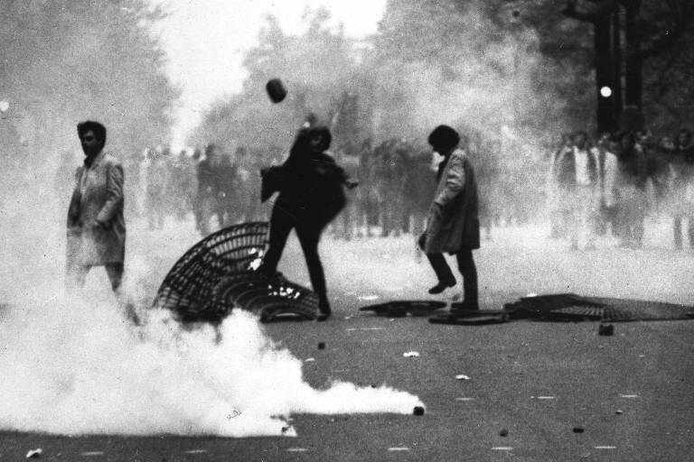 Três homens são vistos em uma avenida de costas lado a lado, sendo que os dois à direita atiram pedras contra um grupo de policiais de tropa de choque à distância; atrás dos manifestantes aparecem uma lata de lixo de ferro retorcida e bombas no chão soltando gás lacrimogêneo