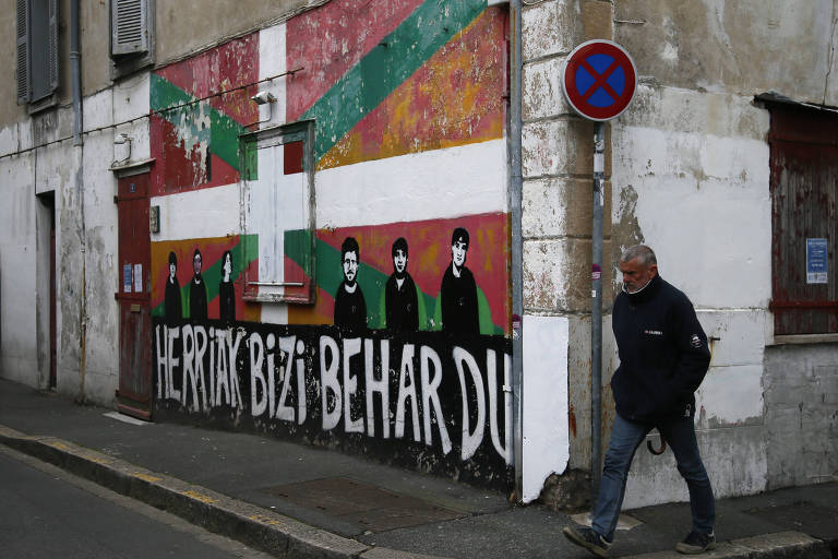 Homem de casaco preto e calça jeans passa por esquina ao lado de mural com a bandeira basca e seis caricaturas de homens, com a expressão 'O povo quer viver' escrita em letra de imprensa branca sob fundo preto
