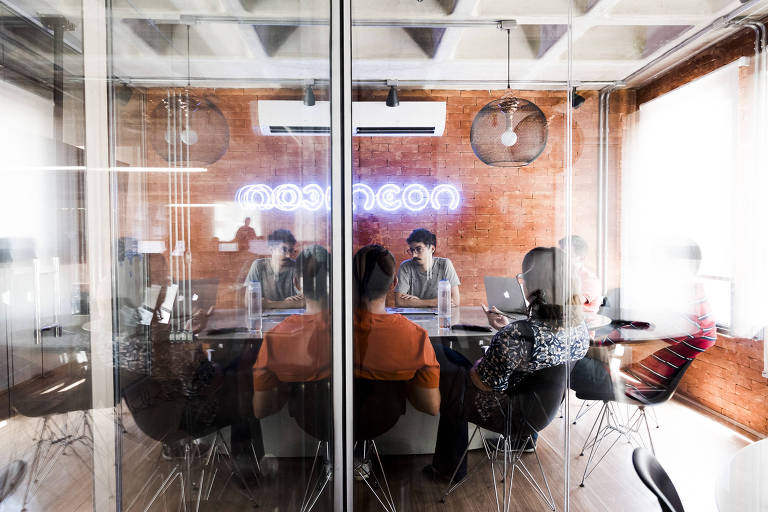 A imagem mostra uma sala de reunião com paredes de vidro. Dentro， há seis pessoas sentadas ao redor de uma mesa retangular. A decoração inclui uma parede de tijolos expostos e um letreiro de neon com a palavra 039;neon039;. A iluminação é suave， e há lâmpadas pendentes no teto.