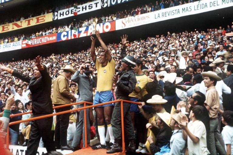Homem com shorts azul e camisa amarela levanta taça em meio a multidão