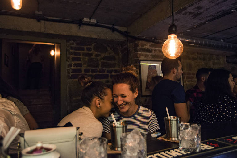 Duas jovens sentadas no balção de um bar, com as cabeças próximas, riem, mostrando intimidade