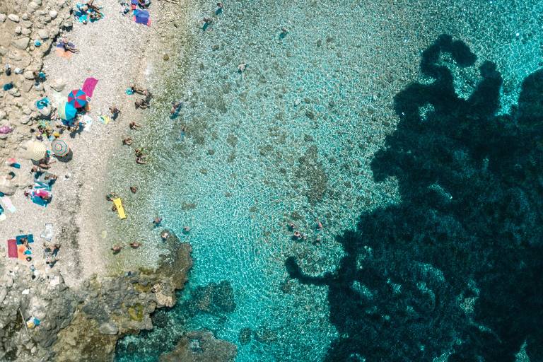 Foto aérea que mostra o mar Mediterrâneo e um pouco da praia. Na praia, há algumas pessoas e guarda-sóis coloridos. O mar tem água turquesa e clara, aparentemente raso