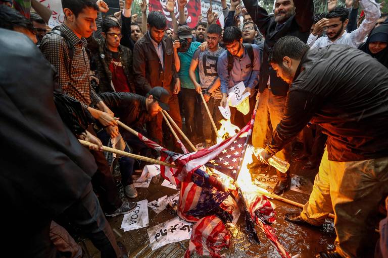 Grupo de dez jovens com bandeiras enroladas em pedaços de madeira as aproximam do fogo para queimá-las