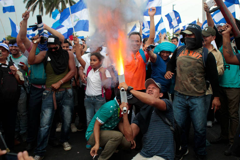 Os fogos são disparados por dois homens, que estão agachados; atrás deles, grupo de manifestantes com bandeiras da Nicarágua, alguns deles mascarados, outros com morteiros similares ao usado para disparar os fogos na mão
