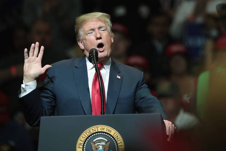 De terno preto, camisa branca e gravata vermelha, Donald Trump levanta a mão enquanto fala em um púlpito com o selo do Presidente dos EUA; ao fundo, apoiadores aparecem desfocados