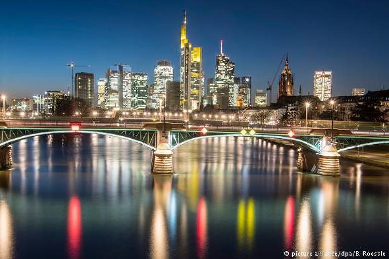 Centro de Frankfurt à noite. Em primeiro plano, o rio Meno, e uma ponte que o cruza. Ao fundo, vários arranha-céus da cidade, com janelas iluminadas.