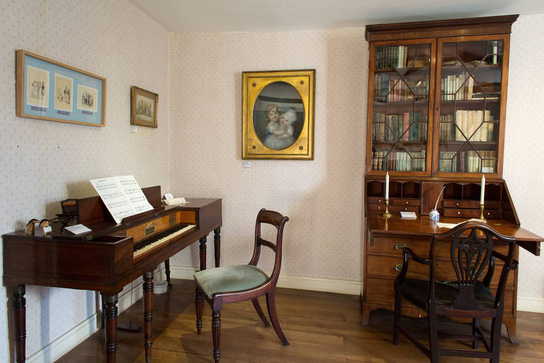 Ambiente do museu de Jane Austen, instalado na casa onde a escritora viveu, em Chawton, Inglaterra; ambiente mostra um cravo e uma estante de livros, além de alguns quadros na parede