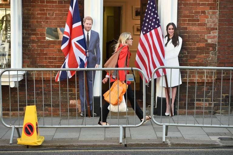 Os displays estão ao lado dos batentes brancos de uma porta de uma casa de tijolos laranjas; à esquerda, aparece o príncipe Harry, em uma foto de terno ao lado de uma bandeira verdadeira do Reino Unido; já Meghan, à direita, veste blazer branco e tem uma bandeira dos Estados Unidos à sua esquerda; a calçada é cercada por duas grades móveis; do lado direito também aparece um cone de sinalização amarelo