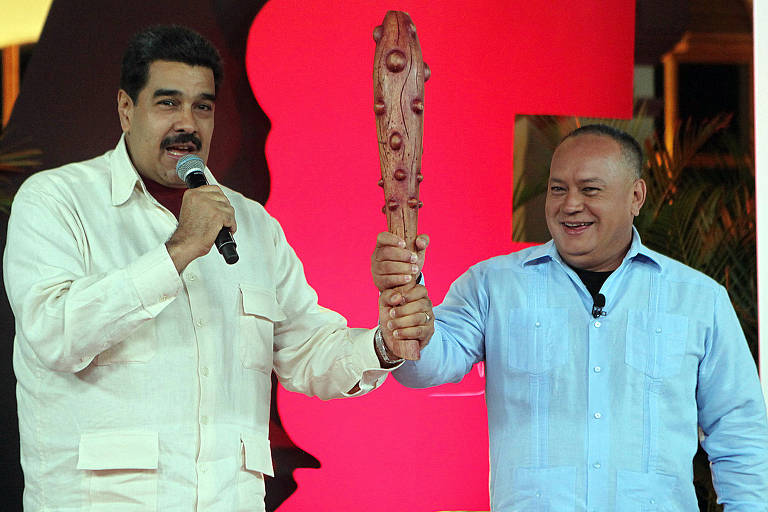 Nicolás Maduro está de camisa amarela, segurando um microfone enquanto fala, e, de camisa azul, Diosdado Cabello aparece sorrindo; o porrete que os dois seguram é marrom e o fundo do cenário, vermelho