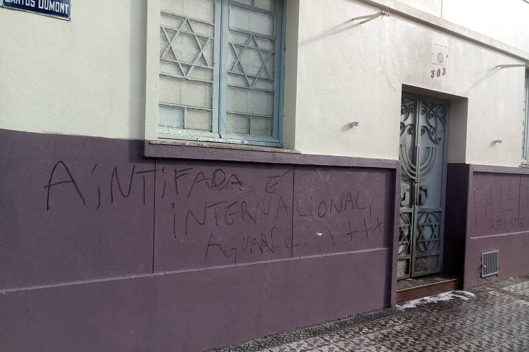 Parede de prédio de muro branco na parte alta e lilás abaixo das janelas, cujas esquadrias são decoradas com a estrela de Davi; na parte lilás, a seguinte expressão em tinta preta: "A Intifada é internacional"