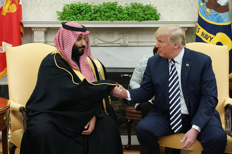 De terno azul marinho, camisa branca e gravata azul marinho e branca listrada na diagonal, Trump aperta a mão de Mohammed bin Salman, vestido de túnica preta e o tradicional lenço de cabeça vermelho e branco da família real saudita; os dois estão sentados à frente da lareira do Salão Oval da Casa Branca