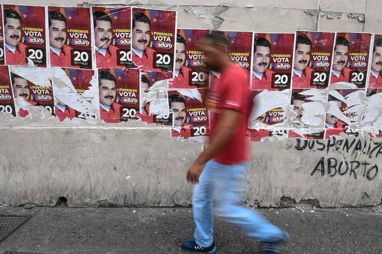 Homem aparece desfocado e traja calça jeans e camiseta vermelha; cartazes de Maduro ocupam parte superior da imagem no muro cinza; abaixo à direita há uma pichação em preto com a expressão "Despenalize o aborto"