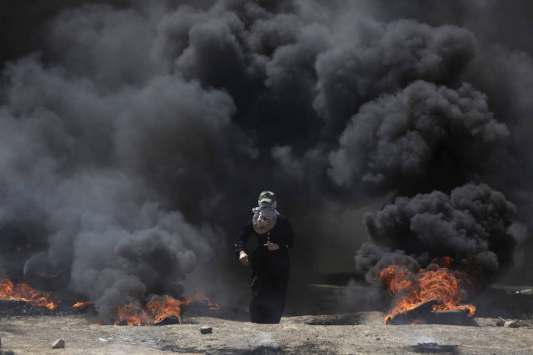 De roupa preta e véu branco, mulher aparece andando entre fumaça preta vinda de pneus queimando