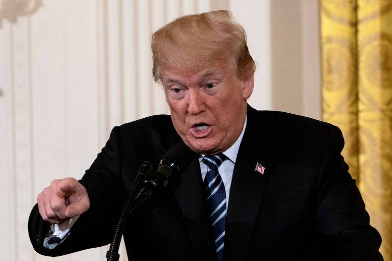 De terno preto, camisa branca e gravata azul e branca listrada, Trump aponta com o dedo direito enquanto fala em um microfone; ao fundo uma parede branca e, à direita, uma cortina dourada
