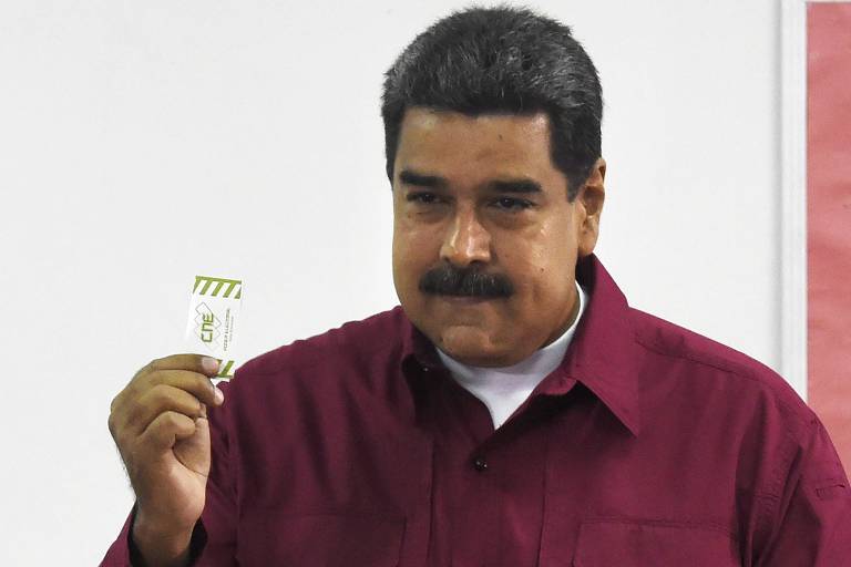 Maduro aparece de camisa vinho e camiseta branca por baixo, levantando a mão esquerda com um papel entre os dedos; ao fundo, uma parede branca
