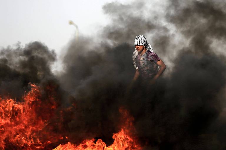 Com lenço quadriculado preto e branco amarrado na cabeça e camiseta roxa, jovem palestino passa por nuvem de fumaça provocada por pneus queimando, que aparecem na parte de baixo da imagem