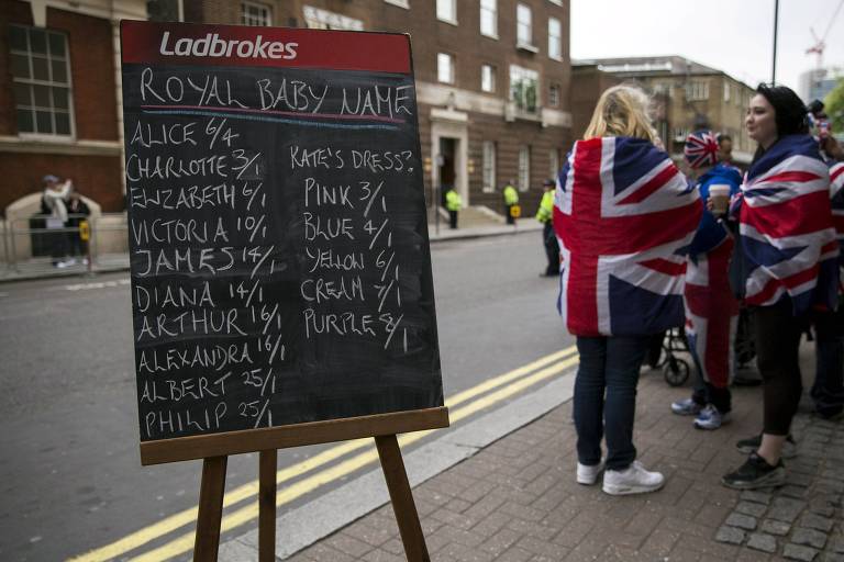 Quadro-negro aparece em pedestal em calçada, com o logo da marca Ladbrokes, de apostas, na parte de cima em fundo vermelho; à direita mulheres enroladas em bandeiras britânicas esperam