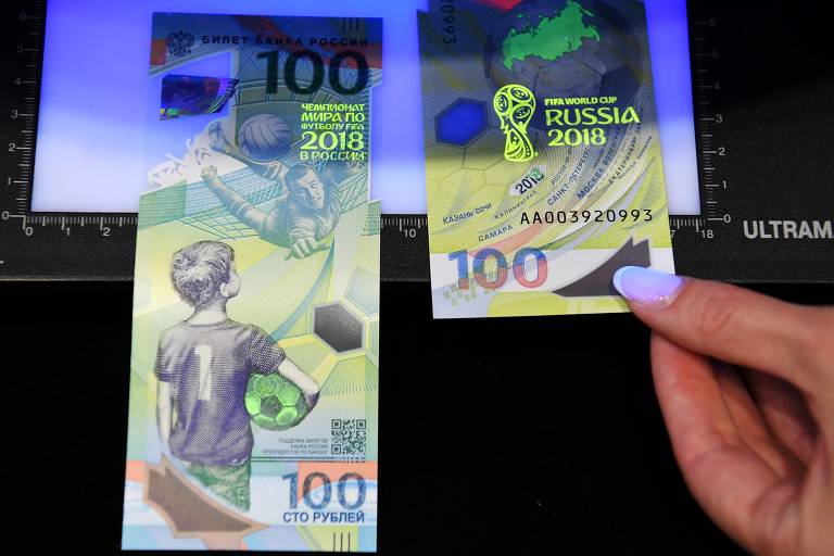 Funcionária do BC russo demonstra itens de segurança da nova nota de 100 rublos, com hologramas e homenageando a Copa