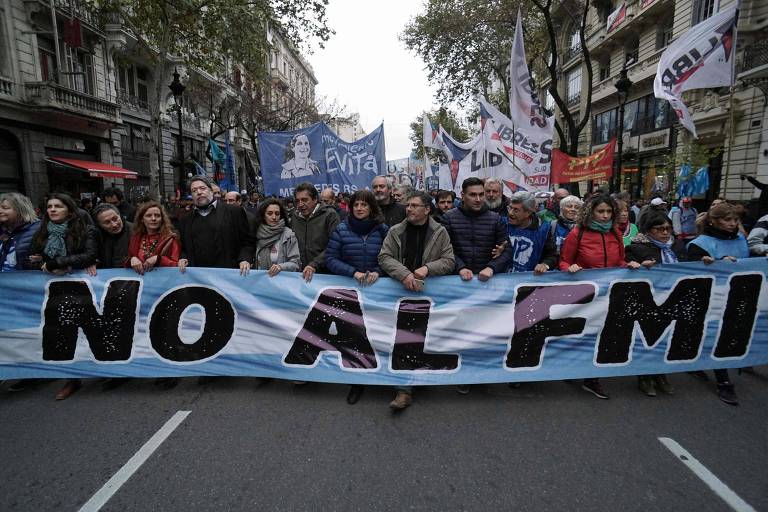 Grupo de dez pessoas carrega faixa com as cores da bandeira argentina e a expressão "não ao FMI" em espanhol em uma rua da capital argentina. Ao fundo, outras pessoas carregam bandeiras de diferentes tamanhos e mensagens políticas