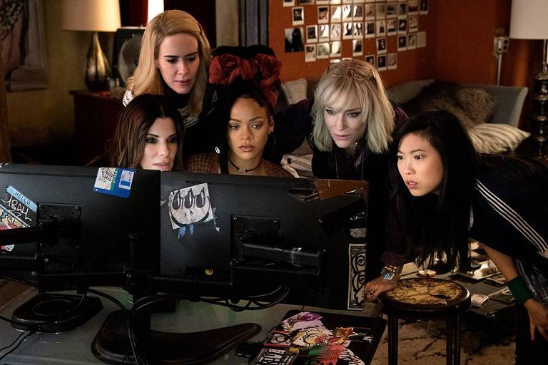 Cinco mulheres ( Sandra Bullock, Sarah Paulson, Rihanna, Cate Blanchett e Awkwafina) olham com rostos concentrados e surpresos para um computador, cuja tela as ilumina; a cena é do filme "Oito Mulheres e Um Segredo"