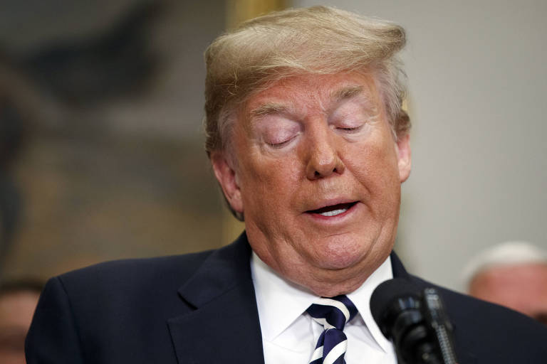 Com terno azul marinho, camisa branca e gravata listrada azul marinho e branca, Trump aparece com os olhos fechados enquanto fala em um microfone à sua frente; ao fundo, desfocada, aparece um pedaço de uma tela