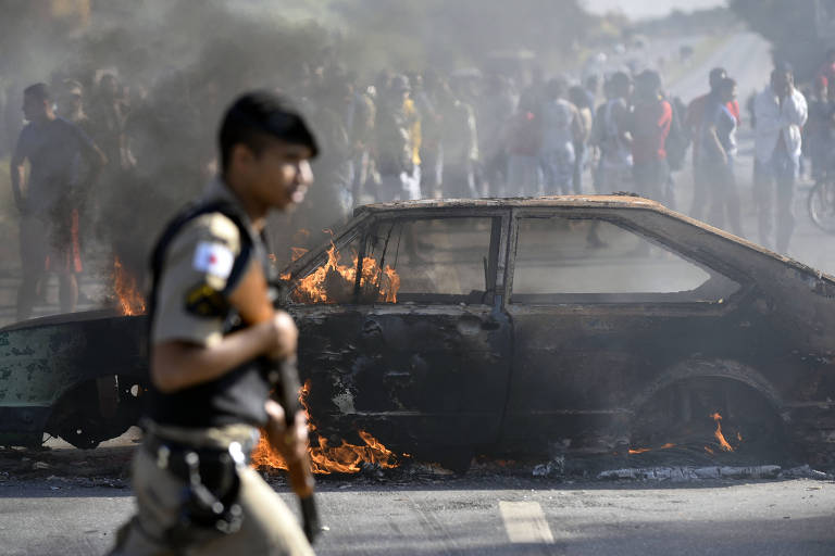 Policial em frente a carro incendiado por manifestantes em Ribeirão das Neves, área metropolitana de Belo Horizonte