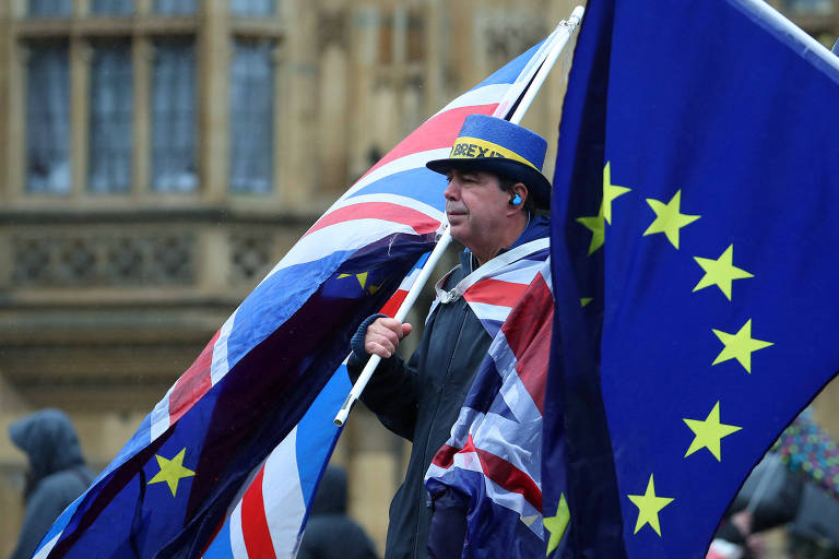 Homem usa chapéu azul com fita amarela, camisa com as cores do Reino Unido e leva em suas mãos uma bandeira britânica e outra da União Europeia; ao lado direito aparece outra bandeira europeia