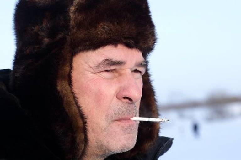 Com neve ao fundo da imagem, homem com roupas de frio tem cigarro em sua boca