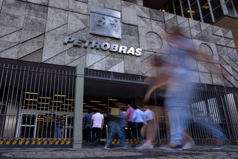 Vista da entrada principal da sede da Petrobras no Rio de Janeiro, onde se lê o logo com o nome da empresa. Pedestres passam na calçada em frente ao prédio