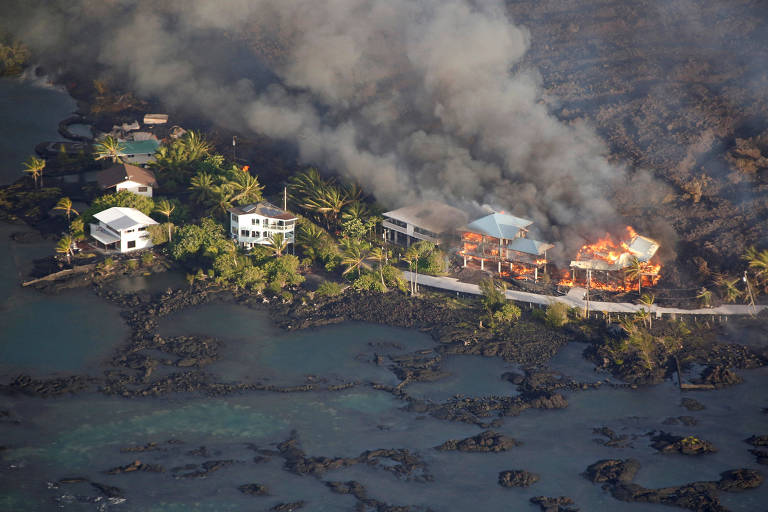 Cindo casas na beira da praia, com lava atrás e duas delas pegando fogo 
