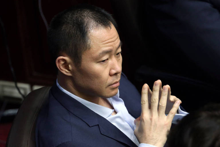 Kenji Fujimori usa blazer azul marinho, camisa branca e une os dedos das mãos enquanto está sentado observando a sessão plenária. Ele é fotografado de cima, aparecendo apenas seu torso.