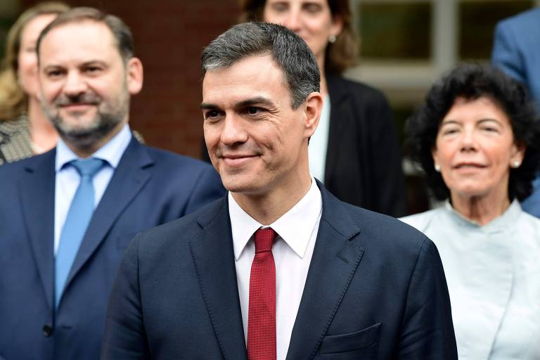 Sánchez usa terno preto, camisa branca e gravata vermelha. Ele aparece sorrindo, com sua porta-voz, María Isabel Celáa, à direita, e seu ministro de Fomento, José Luis Abalos, à esquerda.