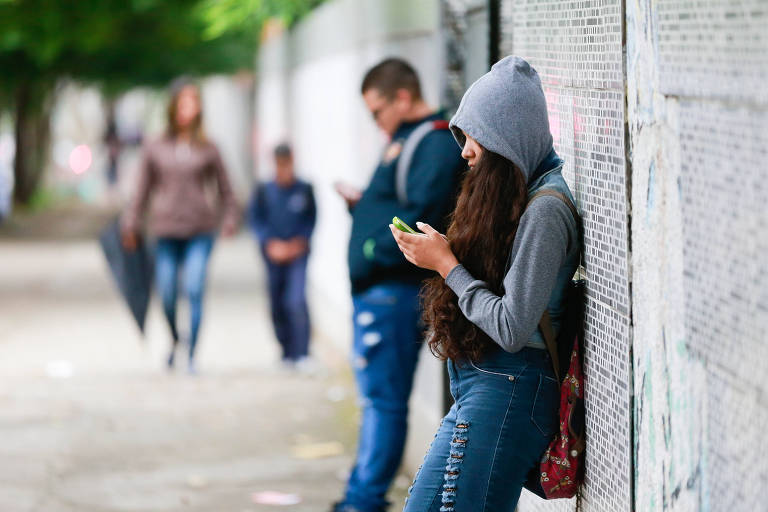 Menina mexe em celular encostada contra muro de escola