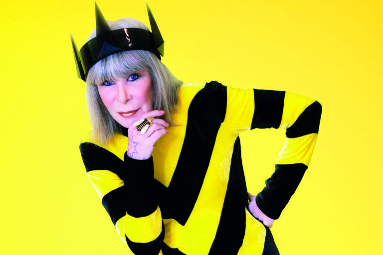 Na imagem, a cantora Rita Lee aparece com um macacão com listras diagonais em preto e amarelo e um chapéu que fazem com que ela pareça uma grande e magra abelha