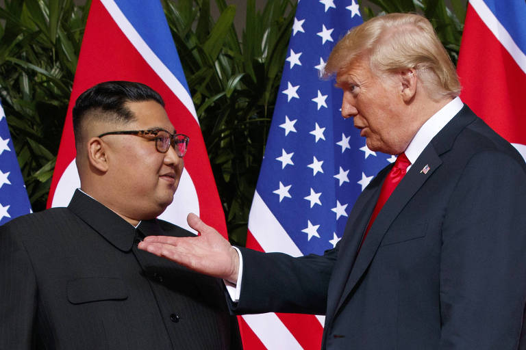 De terno cinza e gravata vermelha, Trump estende a mão enquanto fala com Kim, que usa camisa preta. Ao fundo dos dois, há bandeiras dos Estados Unidos e da Coreia do Norte.